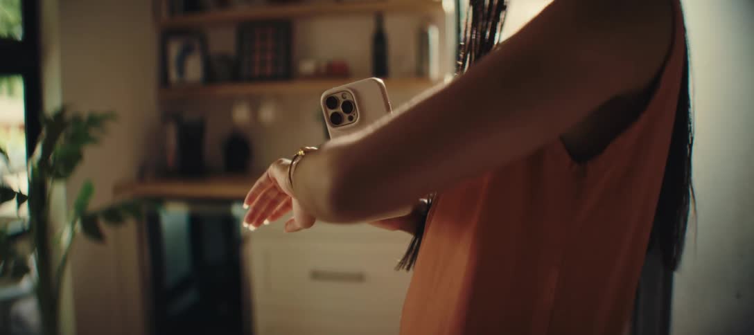 Based on the images from the video a woman is seen standing in a kitchen wearing an orange top and a watch she is holding a smartphone in her hands initially looking at the screen and then turning the phone to face away from her the background featur