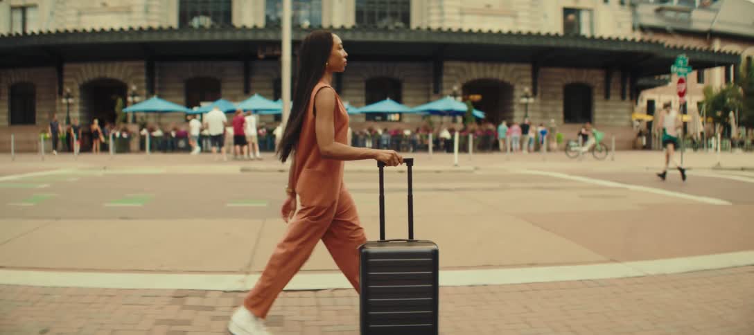 A woman with long dark hair wearing a loose rust colored sleeveless jumpsuit and white shoes is walking with a dark colored rolling suitcase on a sidewalk in a city