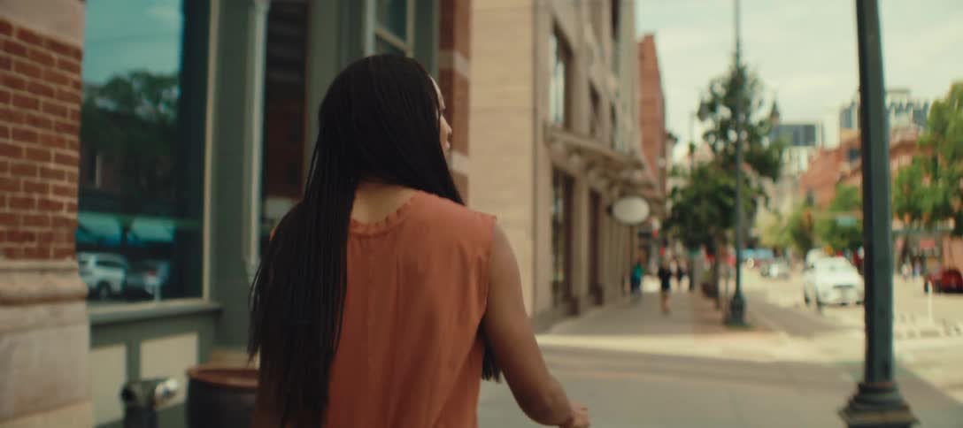 A young woman with braided hair wearing orange clothing walks along the sidewalk at midday rolling her luggage with her