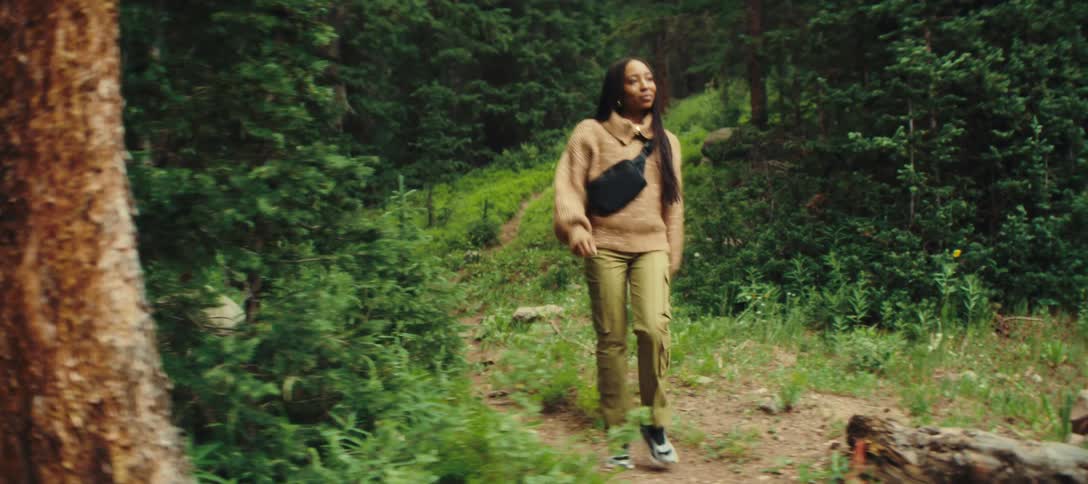 A woman with long dark hair wearing a light brown sweater green pants and a black bag across her chest walks along a narrow dirt path through a dense forest