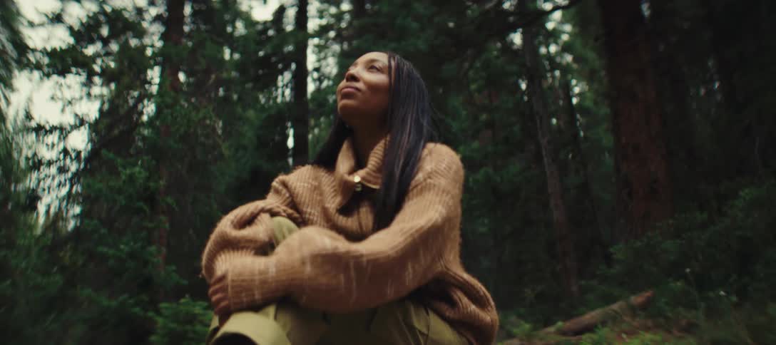 Woman sitting in the forest wearing a brown sweater with a contemplative expression