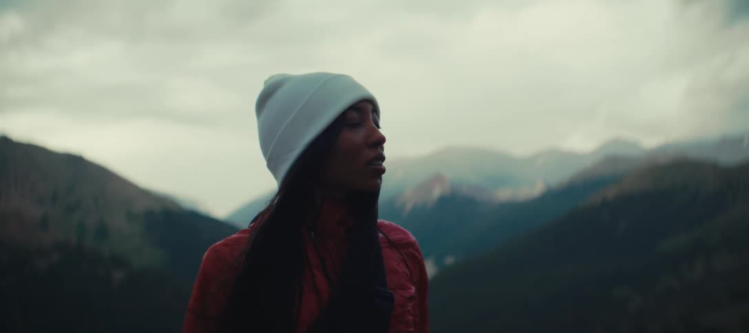 A woman wearing a red jacket and a white beanie stands on a hill looking out at a hazy mountain range under a cloudy sky