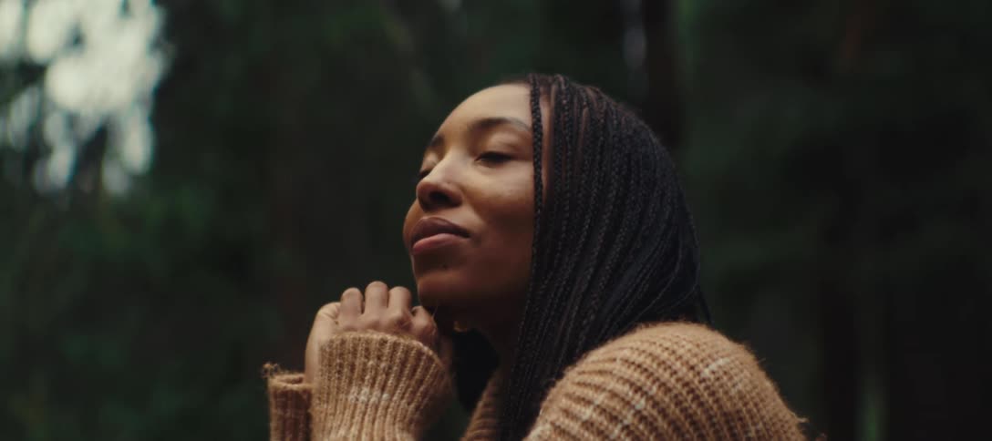 A woman surrounded by trees rests her chin on her hand for a moment as she looks around and parcially smiles