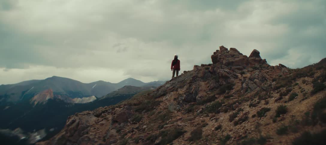 A person wearing a hoodie stands on the peak of a rock on a cloudy day