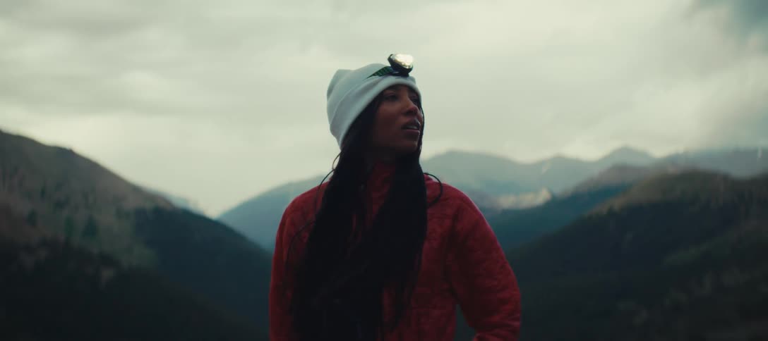 A woman on a mountain range wearing a cap with a lantern looks around and is emotionally moved