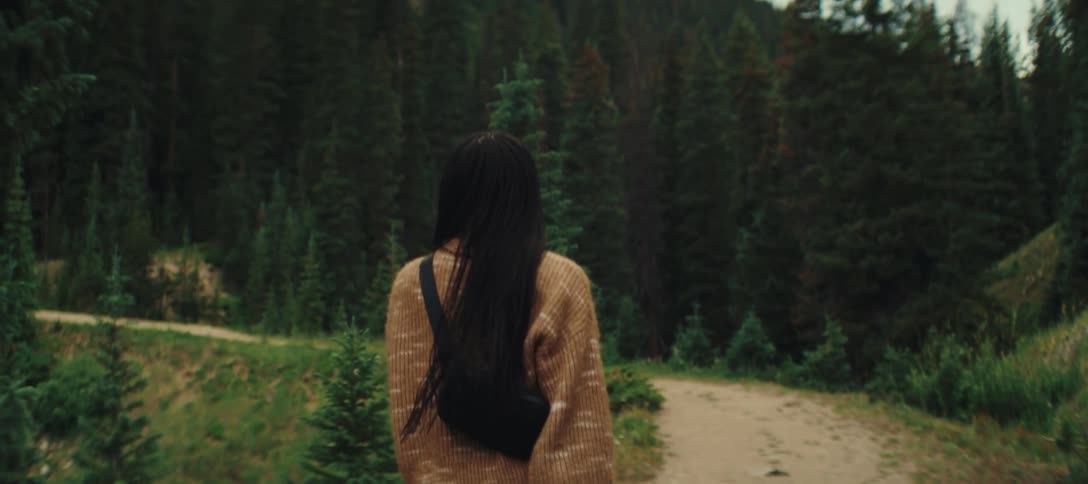 A woman is seen from behind walking away from the camera on a dirt path she has long dark hair and is wearing a tan sweater and green pants
