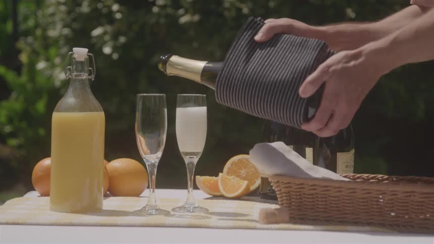 A persons hands pouring champagne from a bottle into a glass on a table which also has a bottle of orange juice oranges and a basket