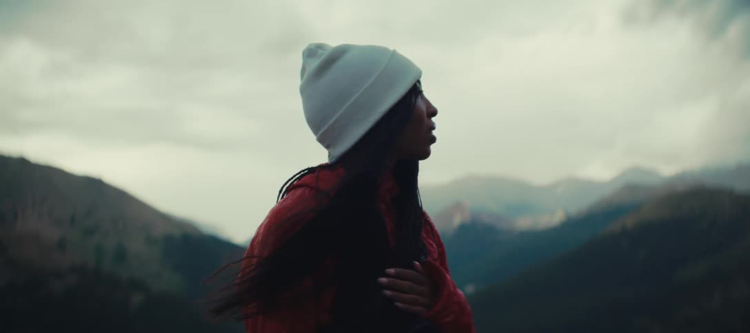 The video shows a woman standing in a mountainous landscape wearing a white beanie and a red jacket she looks up and inhales deeply taking in the fresh air and the vast scenery around her