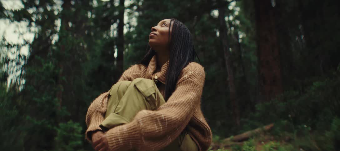 A young woman with long braided hair wearing a brown sweatshirt trousers and sneakers sits on a rock in the woods at midday smiling