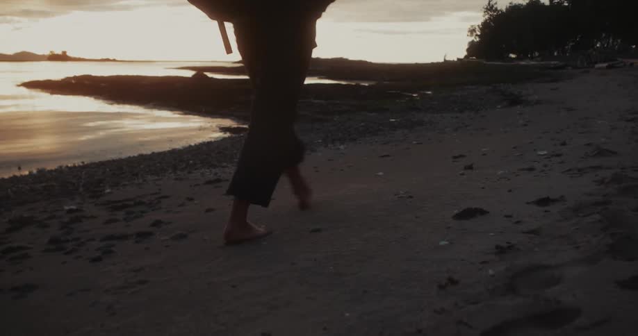 A person walks barefoot on a sandy beach next to the water with the sun setting in the distance