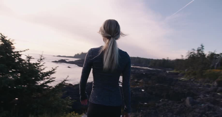 A woman with blonde hair in a ponytail and wearing a long sleeved gray shirt stands on a rocky grassy cliff overlooking a calm ocean with a rocky coastline turning her head to the left as she looks at the water