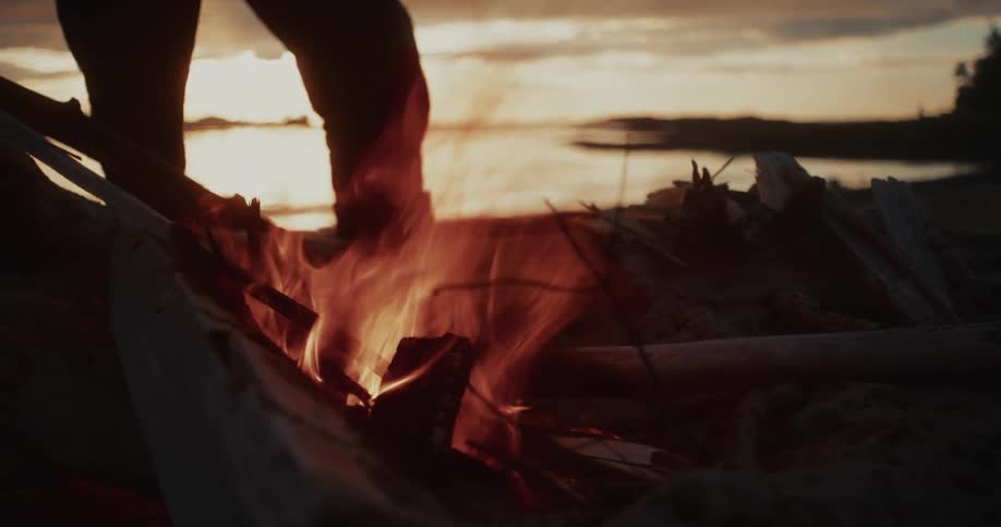 Person standing near a campfire by the lake at sunset