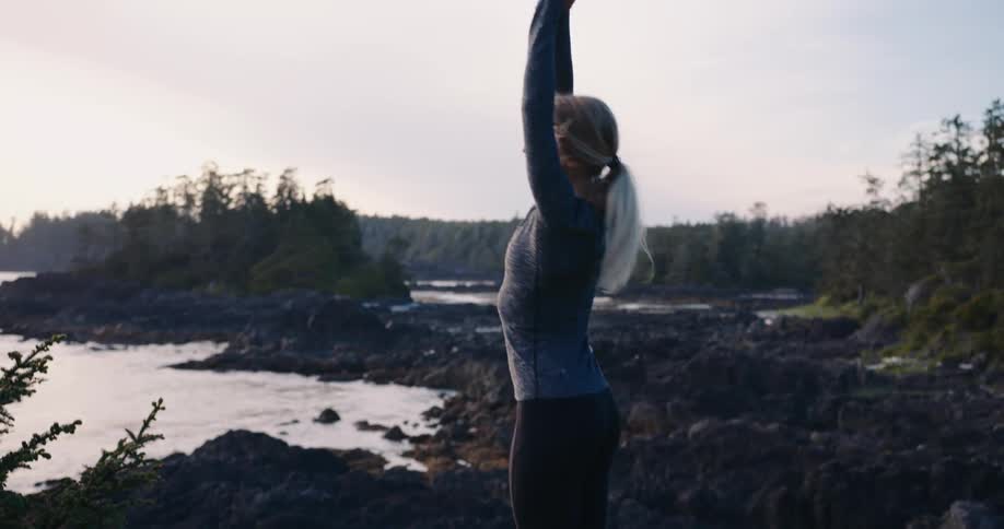 Based on the images from the video a woman with blonde hair is stretching outdoors near a rocky coastline she is wearing dark leggings and a dark blue or gray longsleeved shirt she stretches her leg back with her hand then raises her arms above her h