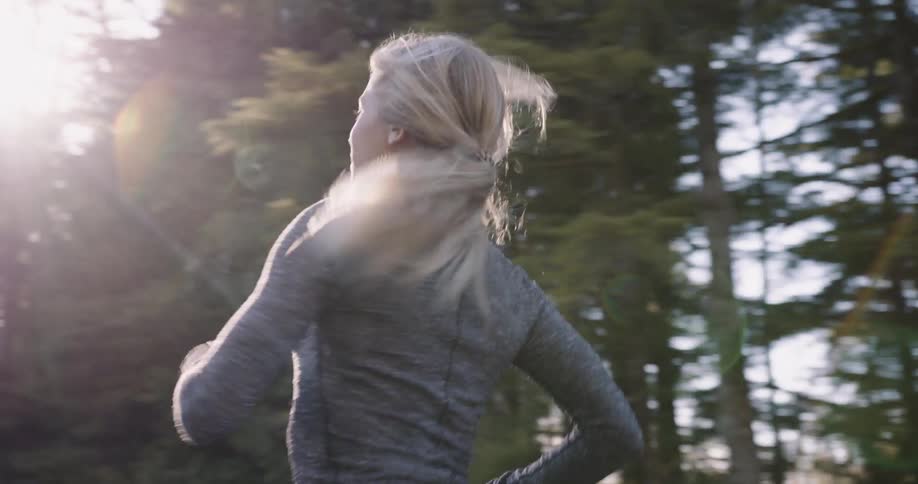 A woman with a ponytail and a gray long sleeved athletic shirt is running away from the camera on a paved path that is surrounded by a thick green forest