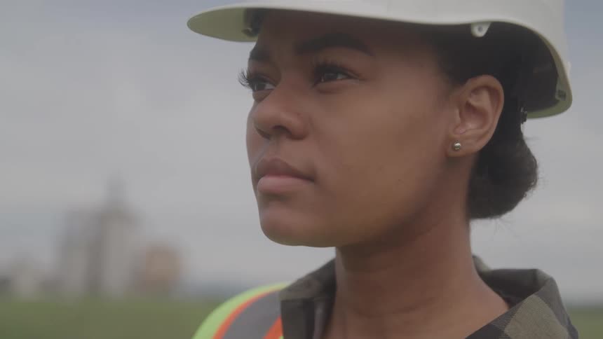A woman in a hardhat and safety vest looking around with a serious expression on her face
