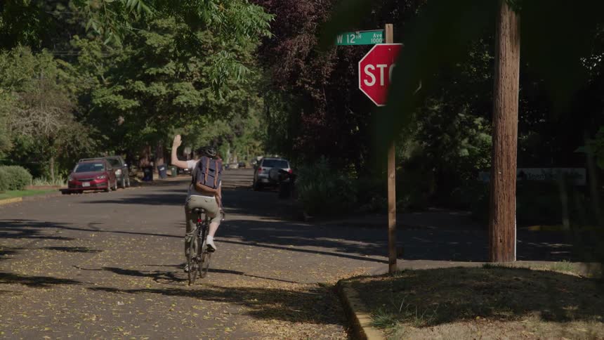 Cyclist with a backpack raises a hand while riding on a treelined street