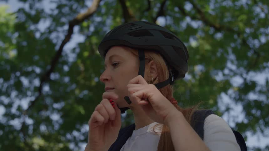 A young woman with long red hair puts on a black bicycle helmet adjusting the straps against a backdrop of green foliage
