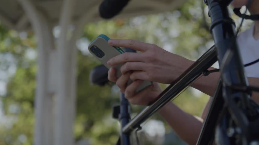A cyclist wearing a helmet sits with her arm resting on her bike while scrolling through her phone in a public park at midday