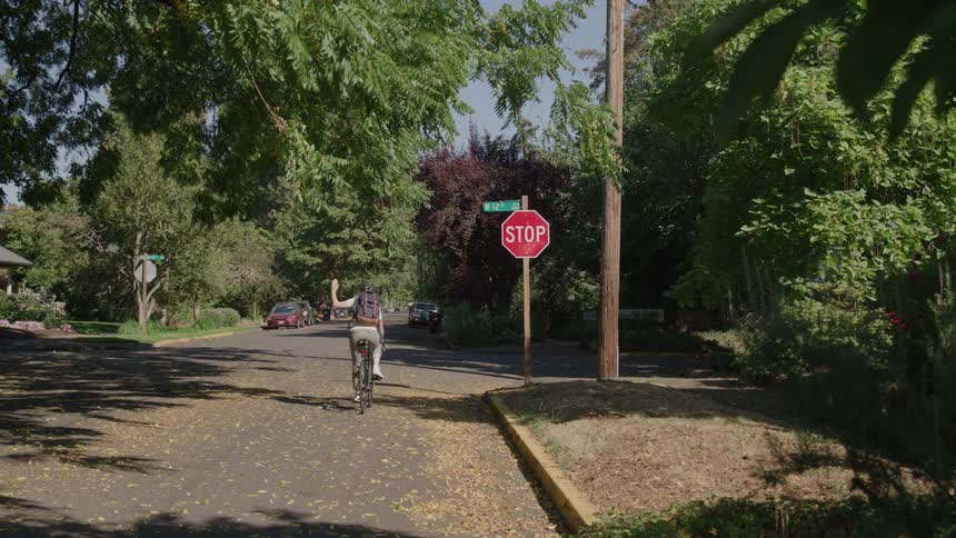 A person with a backpack is riding a bicycle down a treelined residential street and waves at the camera while passing a stop sign