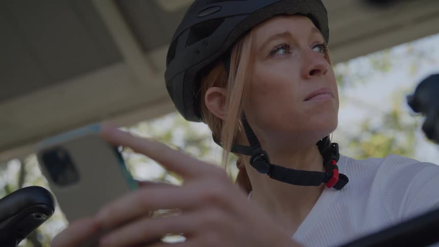 The video shows a closeup of a woman wearing a black bicycle helmet looking intently at her phone she is scrolling on the device while sitting on her bike with a focused and serious expression on her face