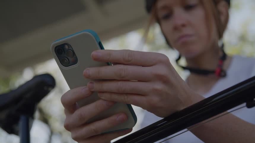 Based on the images from the video a woman wearing a black cycling helmet and a white tshirt is holding a smartphone she appears to be checking the screen while standing next to her bicycle the background consists of blurred trees