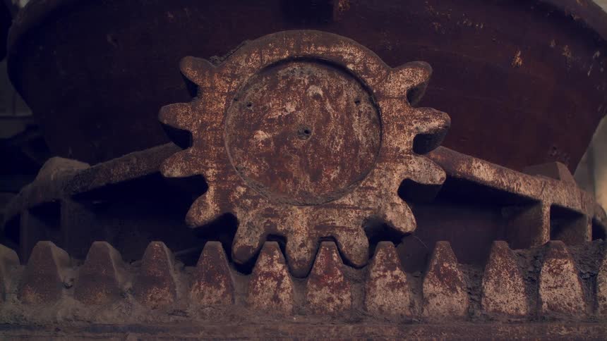 A large rusty metal gear meshing with a stationary rack gear below it