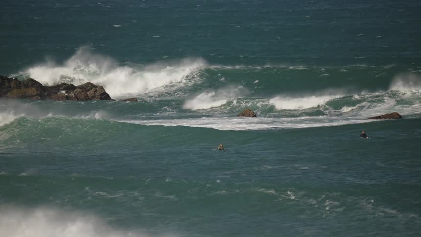 In rough seas a few surfers bob on their boards waiting patiently as large waves crash over rocks in the deep blue ocean