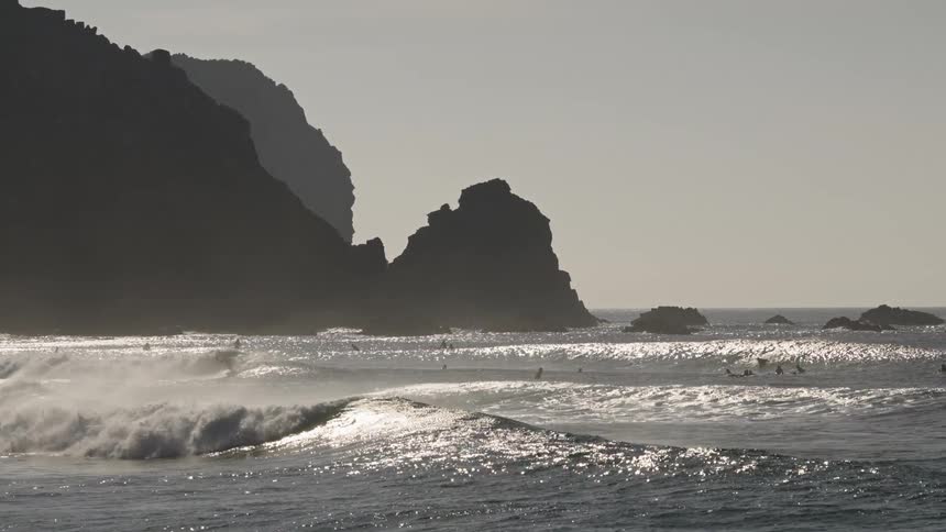 Based on the images from the video the scene shows waves crashing on a body of water with several surfers scattered on the surface in the background there are large dark rocky cliffs and sea stacks silhouetted against a bright hazy sky the overall