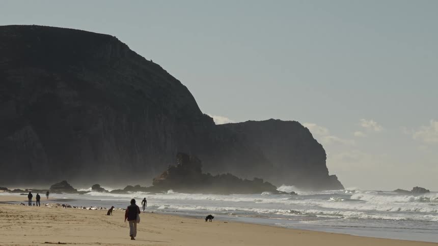 People walking on the sand of a beach with waves crashing and a large rock formation in the background