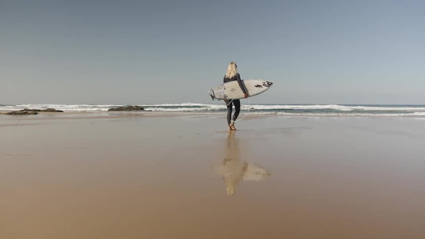Female surfer walking on sand carrying surfboard towards the ocean