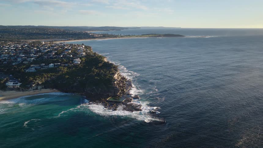 An aerial view of a coastline shows a small town with white houses clustered on a green hill next to a sandy beach with large ocean waves crashing on the rocky shore below as a large rock pool is visible at the bottom of the cliff
