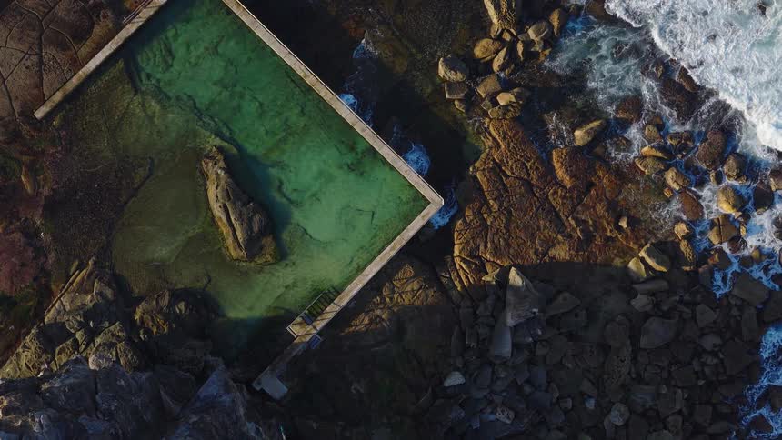 Based on the images from the video the scene is an aerial view of a large rectangular sea pool built into the rocky coastline the water in the pool is a deep green color the pool is surrounded by large rocks and ocean waves can be seen crashing again
