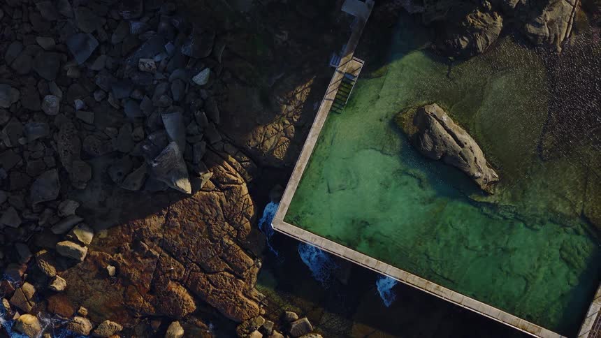 The video shows a highangle shot of a rectangular rock pool next to a rocky shoreline the water in the pool is a clear inviting green contrasting with the dark brown rocks and the blue ocean waves crashing nearby
