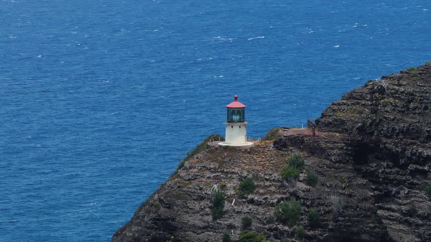 A white lighthouse with a red roof standing prominently on a rugged rocky cliff overlooking a vast deep blue ocean on a clear sunny day