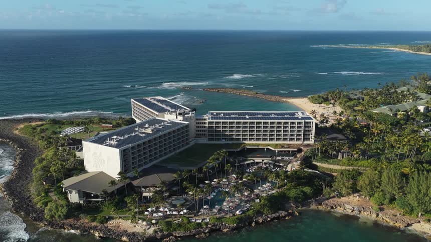 Based on the images from the video the scene is a highangle aerial view of a large modern hotel complex situated on a rocky coastline next to the ocean the hotel has multiple buildings with solar panels on the roof and the grounds feature a pool area
