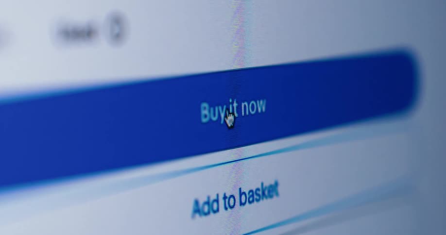 A close shallow focus view shows a computer screen displaying a website with a cursor hovering over a blue button that says buy it now and another button below it that reads add to basket