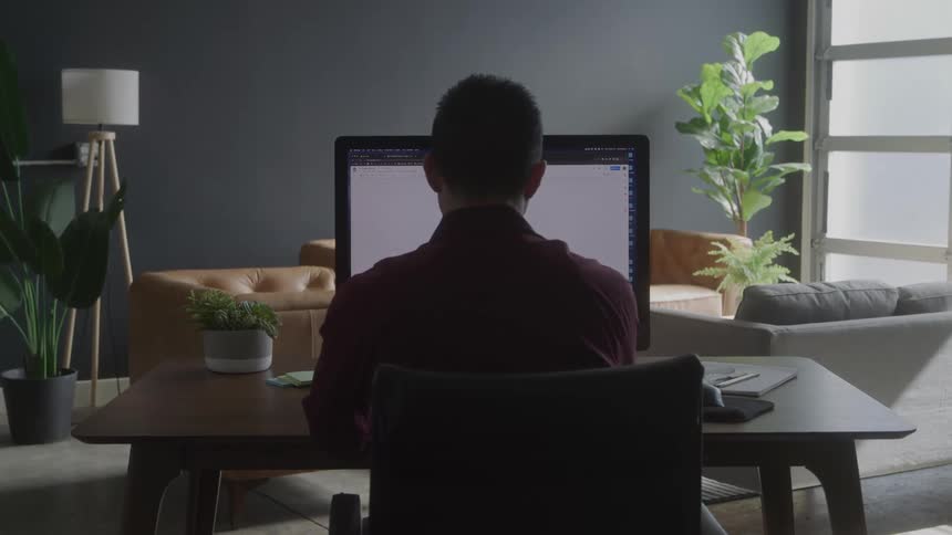 A man sitting at a table in a living room working on a desktop with a large monitor