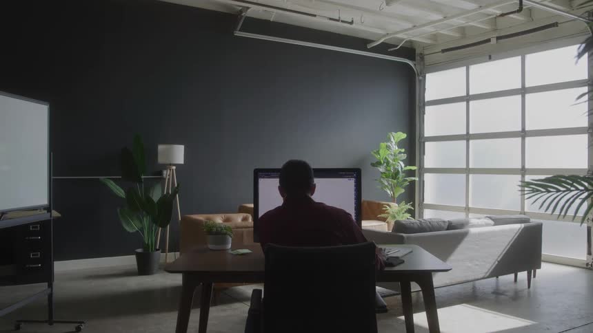 A man wearing a red long sleeved shirt walks up to a desk in a modern office sits down in an office chair and begins to work on a large computer monitor in front of him