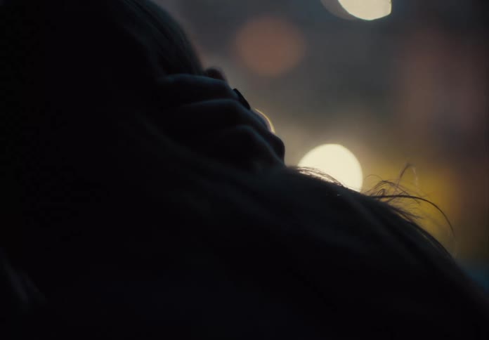 Person in closeup is seen hugging another in the dim light with blurred lights in the background