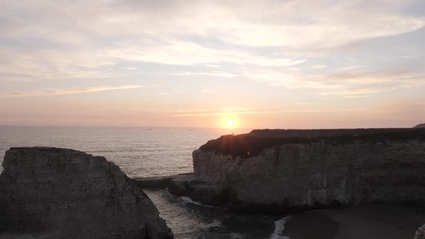 Based on the images from the video the scene is a wide shot of a rocky coastline at what appears to be sunset or sunrise the sun is setting behind a large cliff casting a warm glow on the clouds the waves are gently crashing against the shore and a p