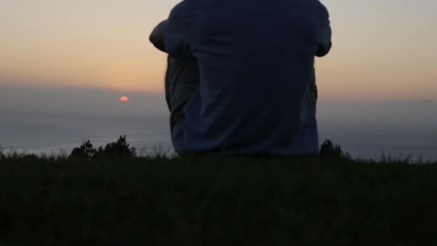 A person sits on a grassy hill facing away from the camera and watches the sun set over a misty landscape