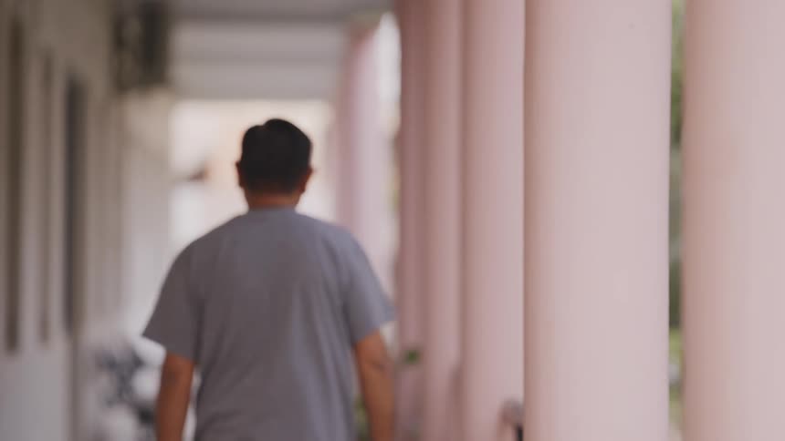 A man with short dark hair is walking away from the camera down a long outdoor hallway lined with large off white pillars on the right side and a building with windows on the left