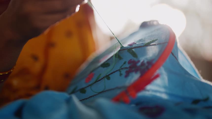 A person is using a needle to pull a green thread through a piece of blue fabric stretched on a red embroidery hoop adding to a pattern of colorful flowers