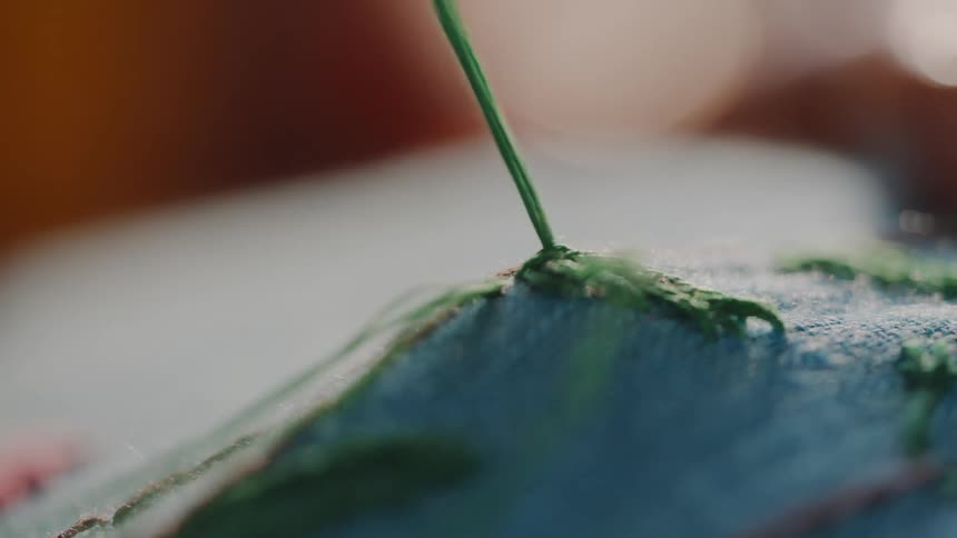 Based on the images from the video the scene is a closeup shot of a persons hands performing an intricate embroidery or sewing technique on a piece of cloth a needle with a green thread is seen pulling through a blue and green pattern creating a text