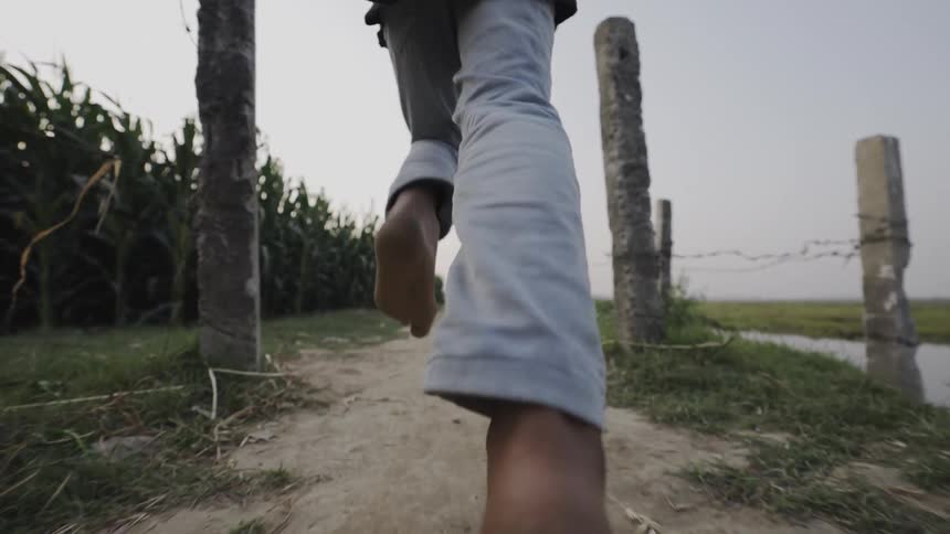 A lowangle shot of a man running barefoot down a dirt path with the camera focused on his feet as he jogs past a cornfield and a body of water