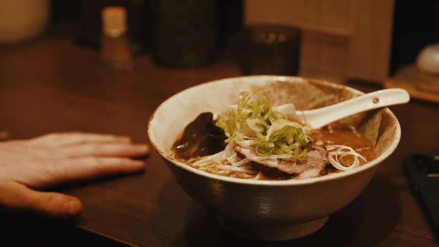 A person is seated at a table with a bowl of steaming asian noodles in front of them