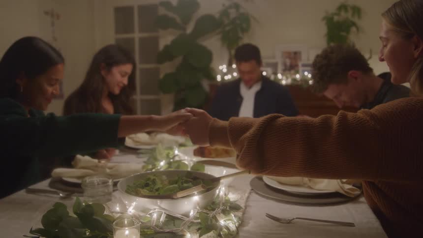 A group of people hold hands and bow their heads in prayer at a dinner table