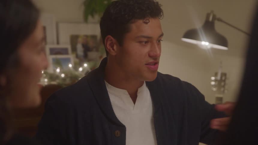 A group of friends having dinner indoors as a young man passes a bowl of vegetables to a young woman while another person hands him a bowl of chicken
