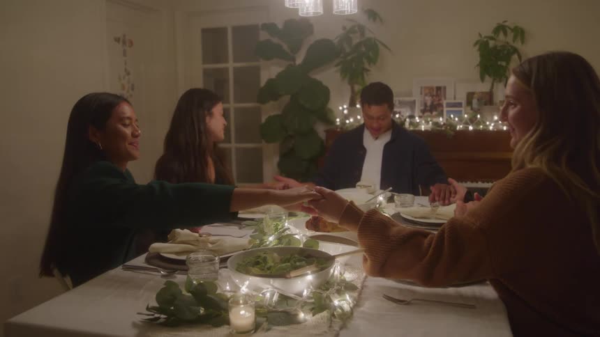 A group of friends hold hands in prayer around a dinner table