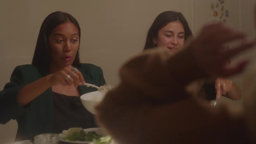 Two women smile and pass a serving bowl at dinner